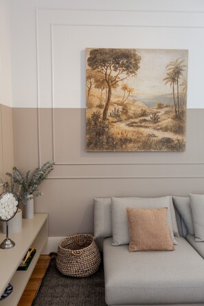 Ein Wohnzimmer mit grauem Sofa, einem gerahmten Landschaftsbild, einem kleinen Tisch mit Lampe und einem gewebten Korb.