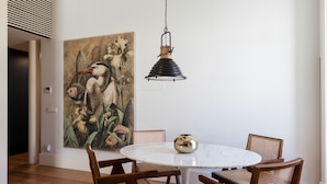 A dining area with a round white table, wooden chairs, a hanging black lamp, and a large floral painting on the wall. - FLH Amazing Praça Comércio Design Flat (Lisbon)