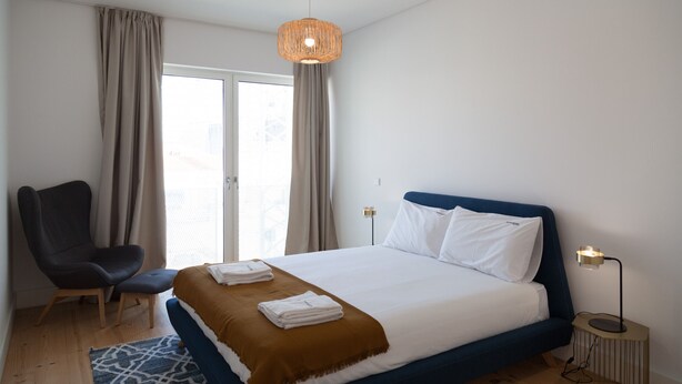 Une chambre d’hôtel moderne avec un grand lit, une chaise, une table de chevet avec une lampe et une vue par la fenêtre, qui est dotée de rideaux.