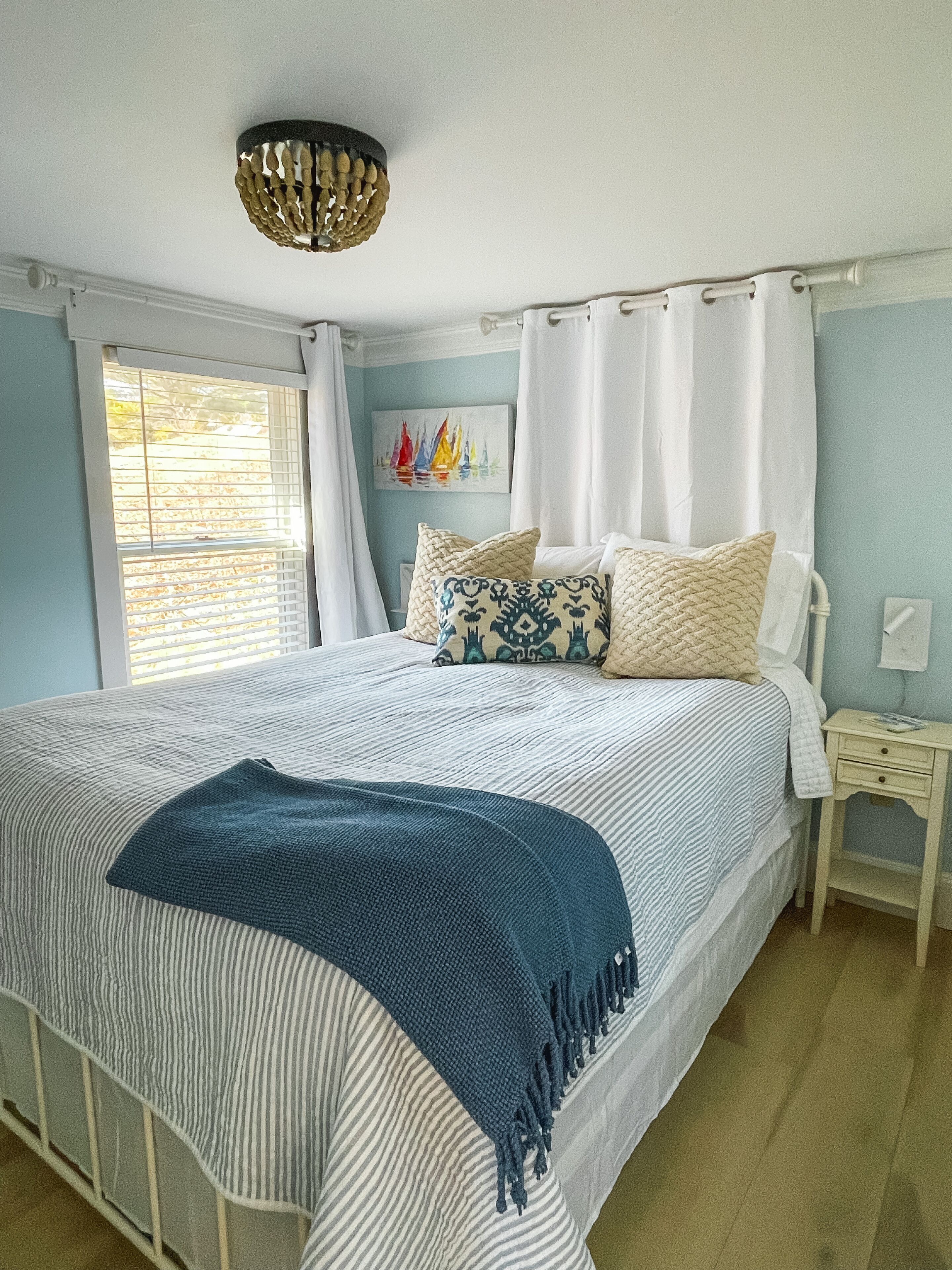 A bedroom with a bed, a bedside table, a nightstand, a window with blinds, and a ceiling light fixture.