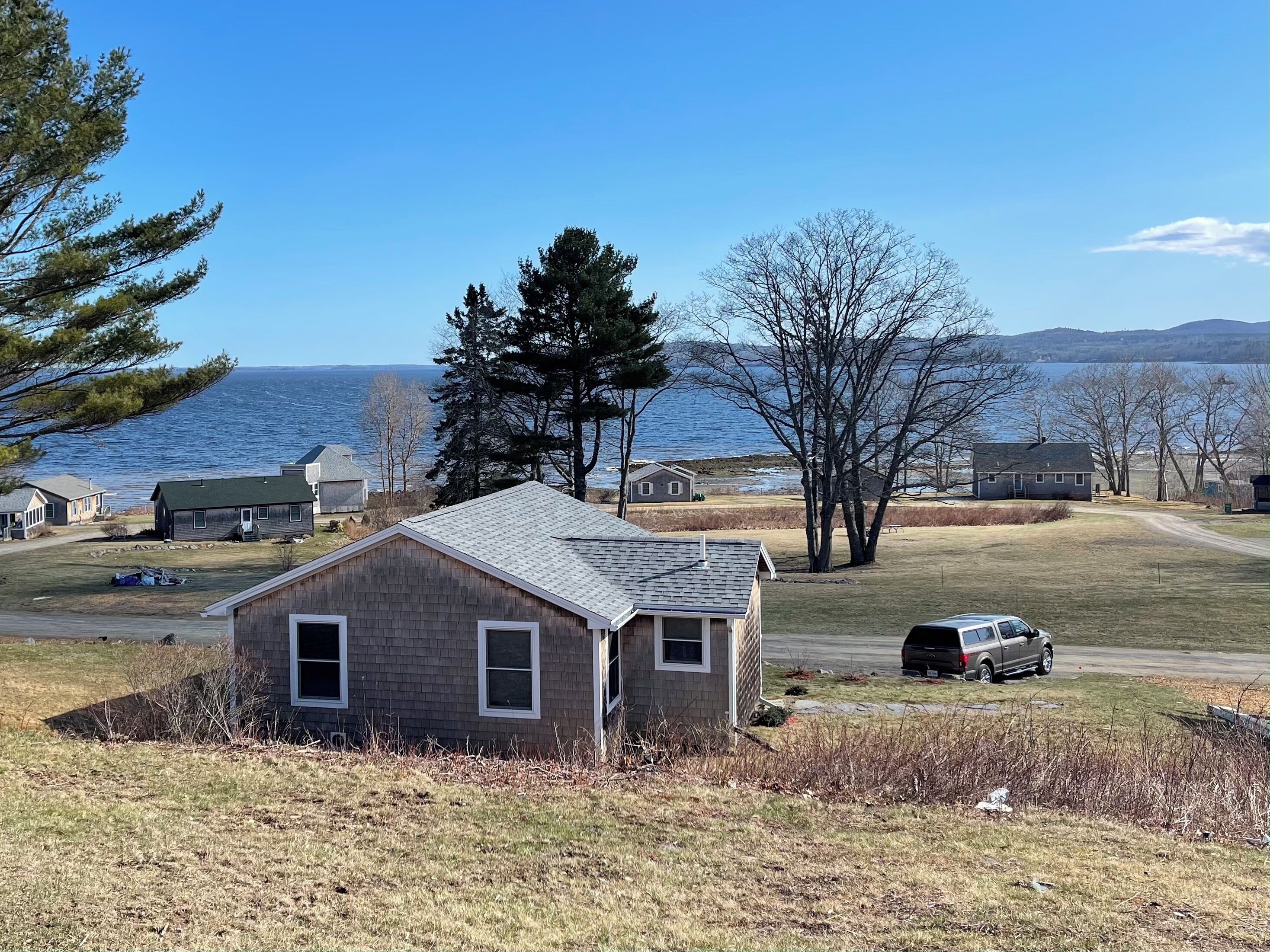 A small house with a sloped roof and a front porch, situated on a grassy area with a view of a body of water and hills in the distance.