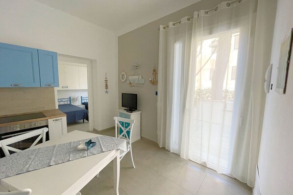 A small, bright room with a kitchenette, a bed, a TV, and a window with curtains.