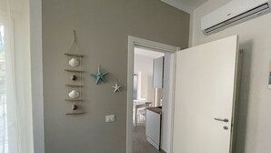 A hallway with a white door, a wall-mounted shelf with decorative items, and a visible kitchen area in the background. - Apartment Giglio near the sea (Marina di Grosseto)