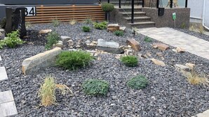 Un edificio moderno con fachada de piedra, una banca de madera y un jardín ajardinado con grava y plantas.