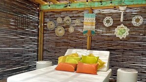 Una cama con colchón blanco y almohadas coloridas, un muro de bambú tejido y adornos colgantes en la pared.