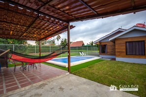 A covered patio with a hammock, a pool, and a wooden house.