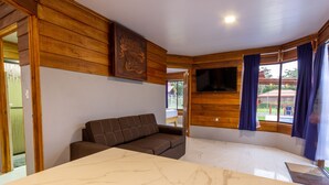 A room with a wooden wall, a brown sofa, a flat-screen TV mounted on the wall, and a window with curtains. - Sunset Arenal House (La Fortuna)