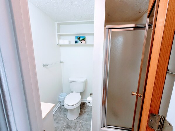 Un baño con inodoro, cabina de ducha y estante con artículos de aseo.