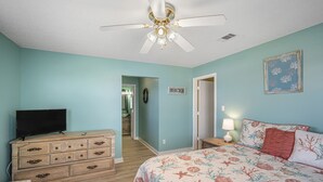 A bedroom with a bed, dresser, television, ceiling fan, and a hallway leading to a bathroom.