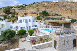 A white building with a pool and outdoor seating, surrounded by greenery and stone structures. - Aegean Gem Mykonos the Most Amazing Sunsets Ever (Mykonos)