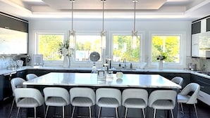 Une cuisine moderne dotée d’un grand îlot central, de tabourets de bar, de luminaires suspendus et de grandes fenêtres offrant une vue sur la verdure.