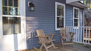 A wooden deck with two rocking chairs and a small table, a white door, and a lantern on the wall. - Aramabad- Close to Beach. Shared Water Access. (Chincoteague)