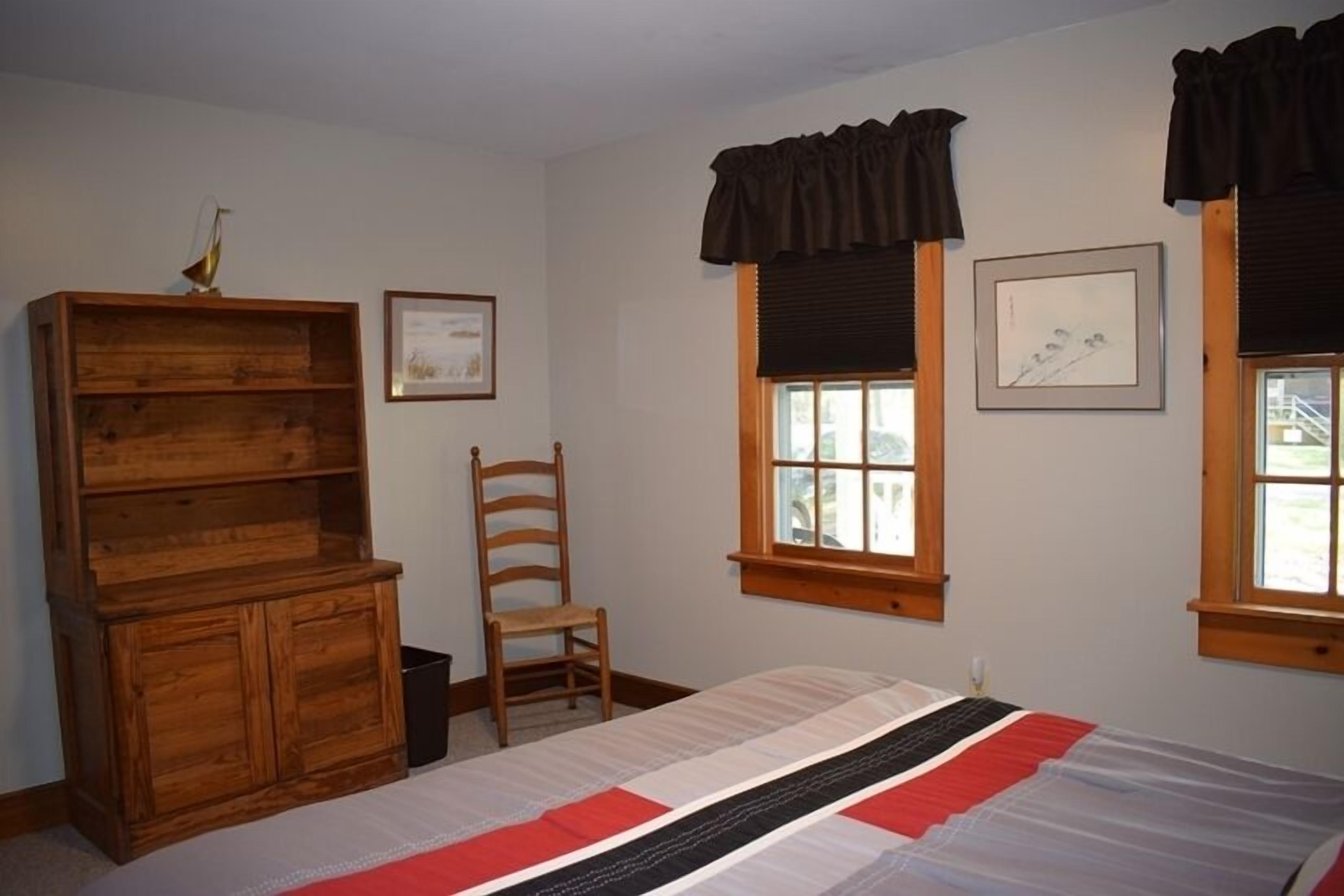 Un dormitorio con cama, cómoda de madera, silla y dos ventanas con cortinas.