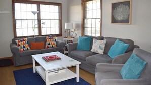 A living room with two grey sofas, a white coffee table, and a blue rug. There are windows with blinds, a lamp, and a framed picture on the wall.