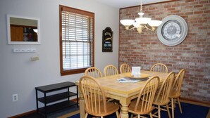 A dining area with a brick wall, a large window with blinds, a chandelier, and a wall clock.