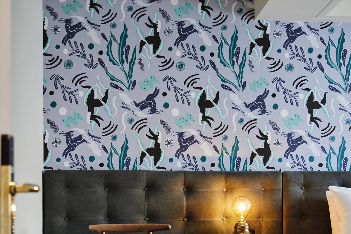 A room with a bed, a desk with a chair, a wall with a patterned design, and a light on the desk.