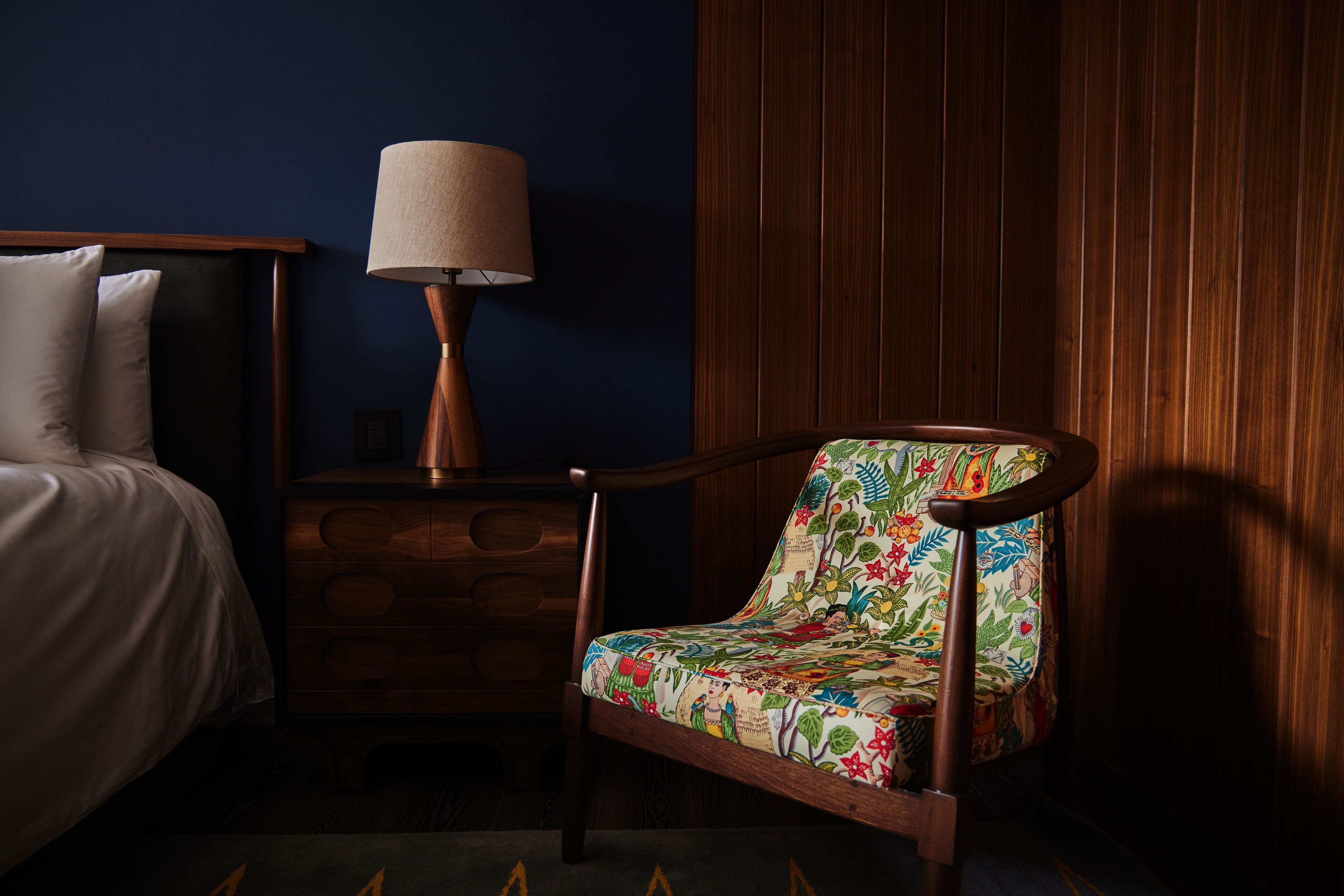 Un dormitorio con cama, mesita de noche, lámpara y una silla con estampado floral.