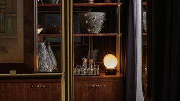Um quarto com iluminação fraca, móveis de madeira, um armário de vidro com objetos decorativos e um abajur aceso.