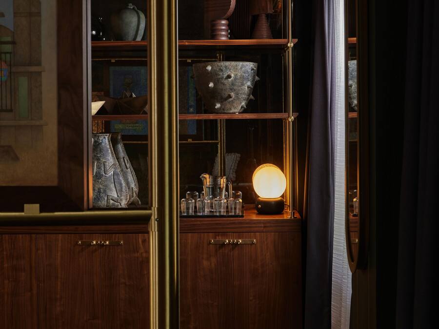 Um quarto com iluminação fraca, móveis de madeira, um armário de vidro com objetos decorativos e um abajur aceso.