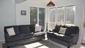 A living room with two grey sofas, a patterned rug, and large windows with a view of trees and a deck. - Avocet- Shared Community Water Access, Close to Beach. (Chincoteague)