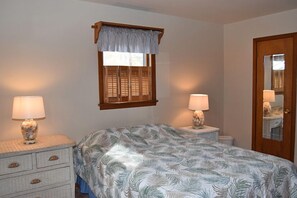 A bedroom with a bed, two bedside tables with lamps, a wooden door, and a window with shutters. - Avocet- Shared Community Water Access, Close to Beach. (Chincoteague)