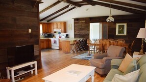 A spacious living area with a television, a white coffee table, and a sofa. A kitchen is visible in the background with wooden cabinets and a dining area with chairs. - Avocet- Shared Community Water Access, Close to Beach. (Chincoteague)