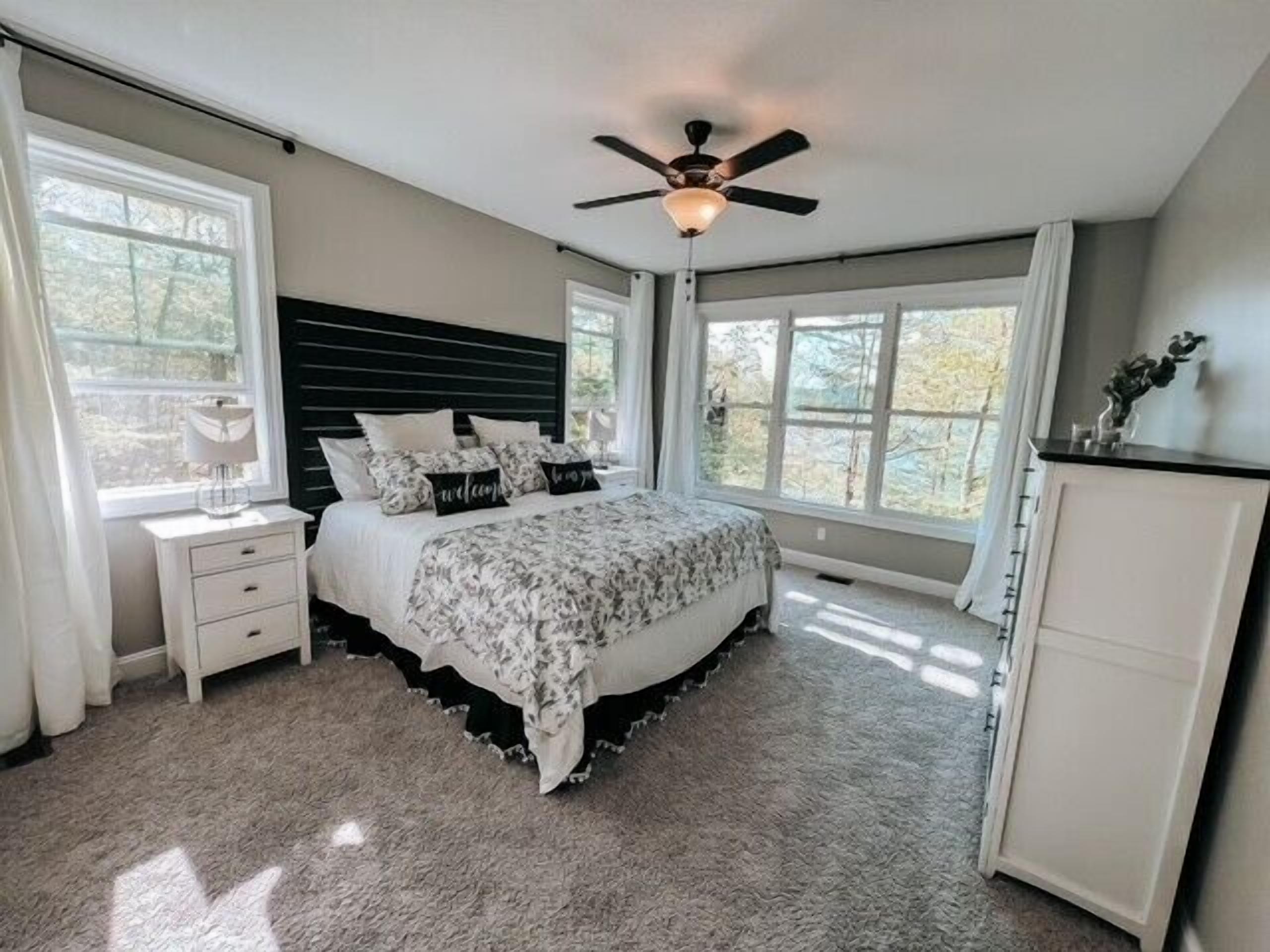 A bedroom with a bed, a nightstand, a dresser, a ceiling fan, and large windows.