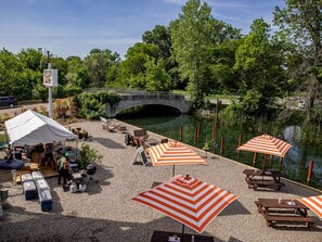 A riverside patio with picnic tables, striped umbrellas, and a covered area with a grill.