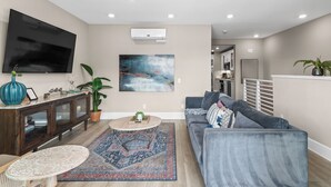 Una sala de estar moderna con un sofá gris, una mesa de centro, un televisor, un cuadro y una cocina al fondo.