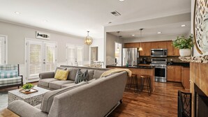 Amplia sala de estar con un sofá gris, piso de madera y una zona de cocina con electrodomésticos modernos.