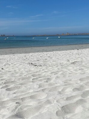 Ein sandiger Strand mit Fußspuren, die zum Wasser führen, Boote in der Ferne und Felsen am Horizont.