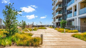 A modern apartment building with a wooden boardwalk leading to the entrance, surrounded by landscaped areas with various plants.