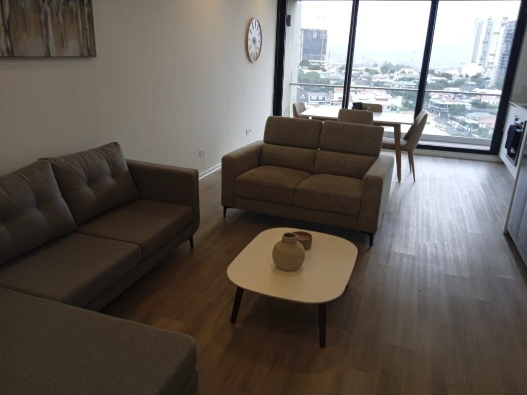 A living room with a sofa, a small table with a vase, and a view of the city through large windows.