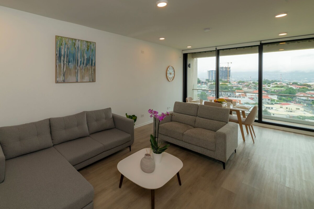 Un salon moderne avec un canapé d’angle, un canapé plus petit, une table basse et une vue sur le paysage urbain grâce à de grandes fenêtres.