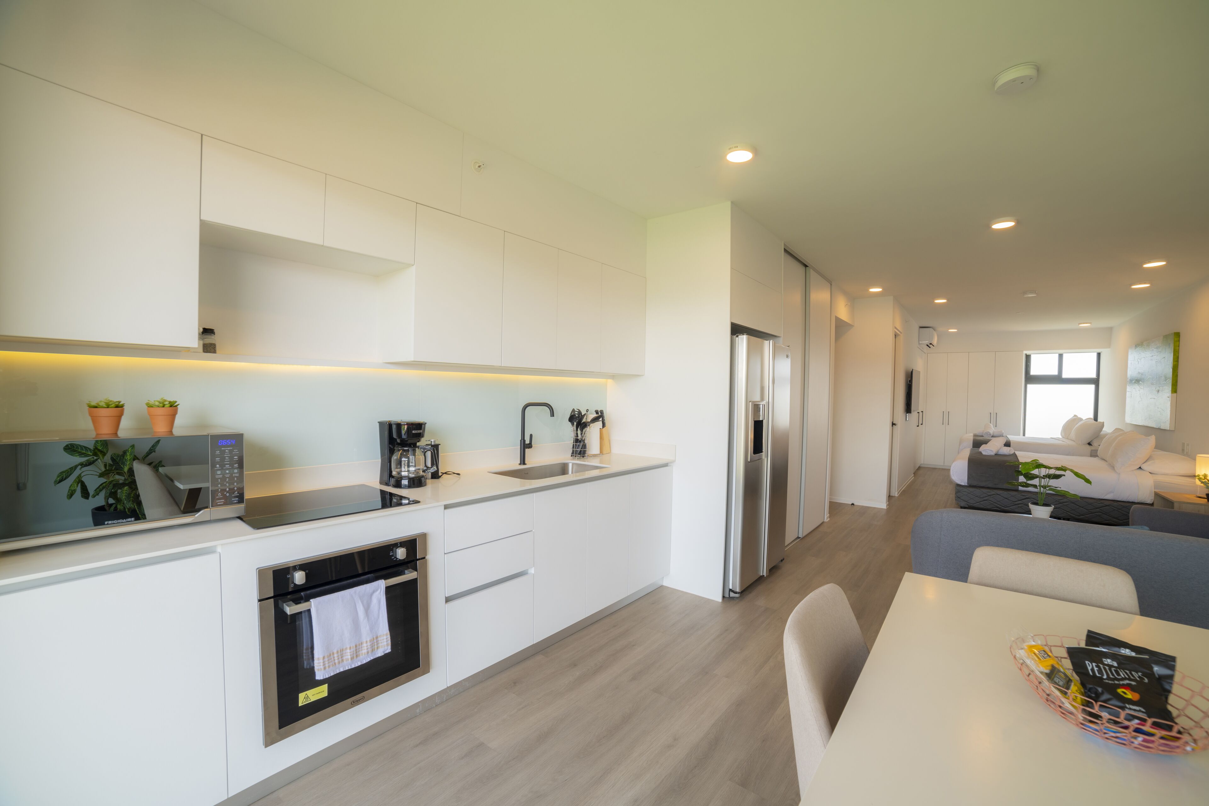 A modern kitchen with built-in appliances, a dining area with a table, and a living space with a sofa and a plant.