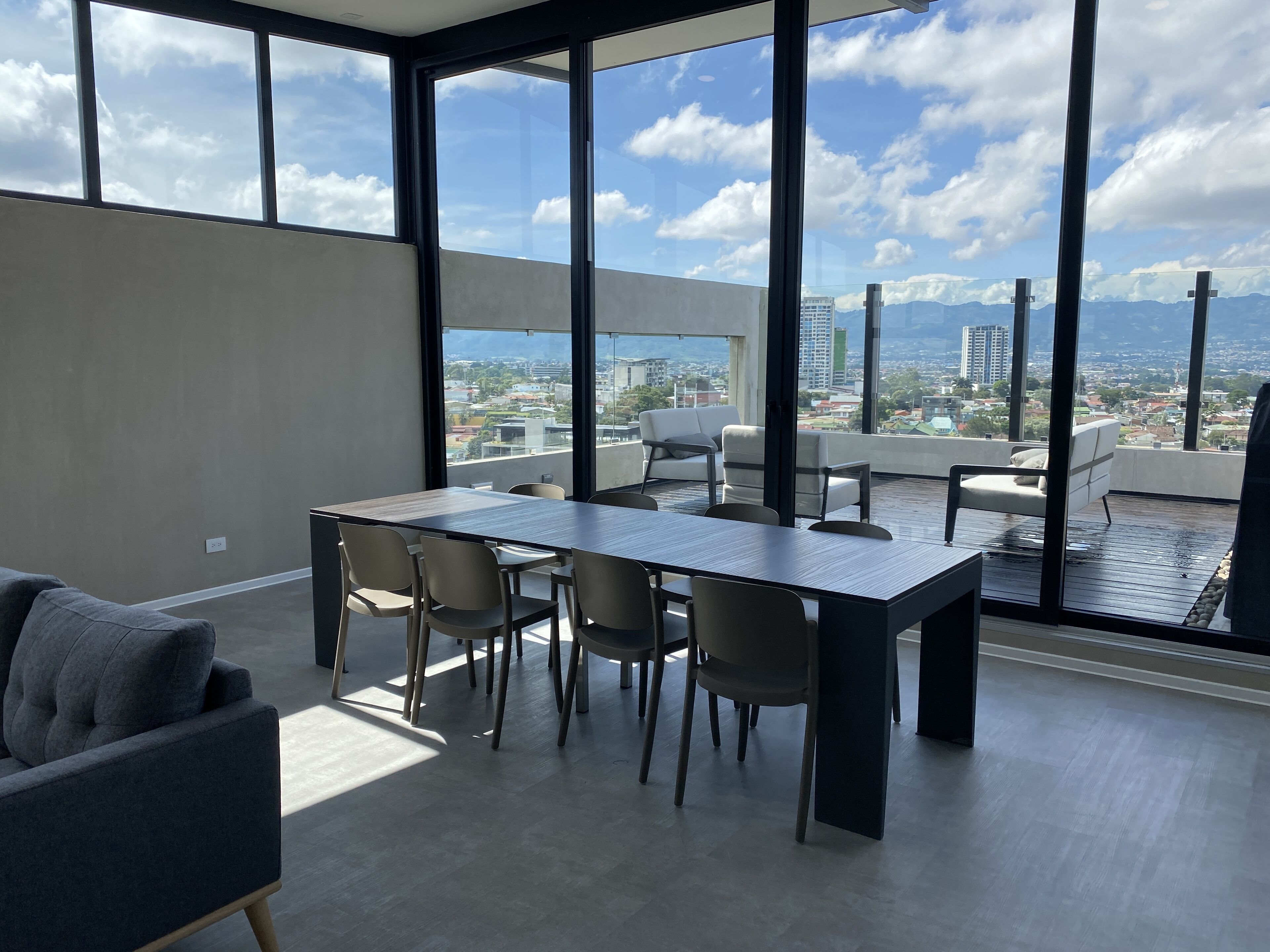 A modern interior with a large dining table and chairs, large windows offering a city view, and a balcony with seating.