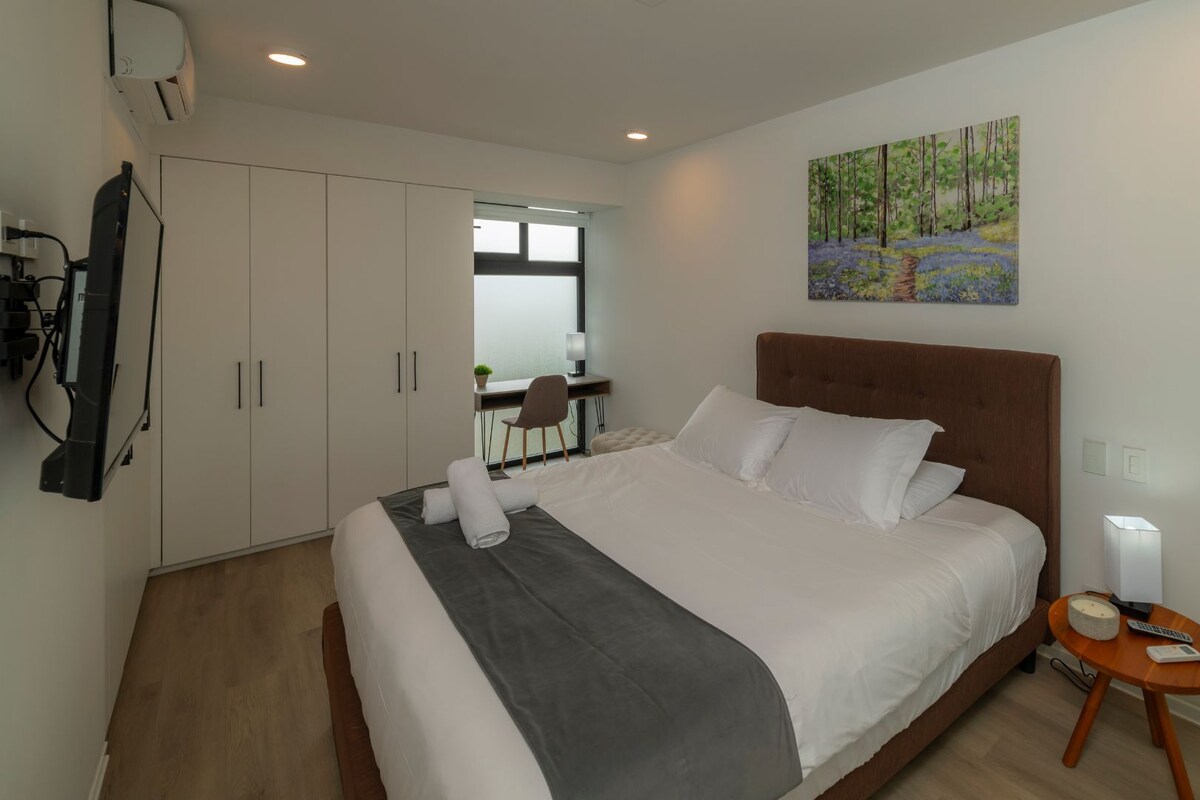 Une chambre d’hôtel avec un grand lit, un bureau avec une chaise, un tableau au mur et une petite table avec une lampe.