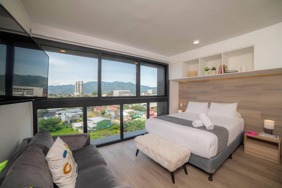 Una habitación de hotel moderna con un ventanal grande, una cama, un sofá y una mesita de noche.