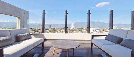 A modern outdoor seating area with a round table, two sofas, and a view of a cityscape and mountains.