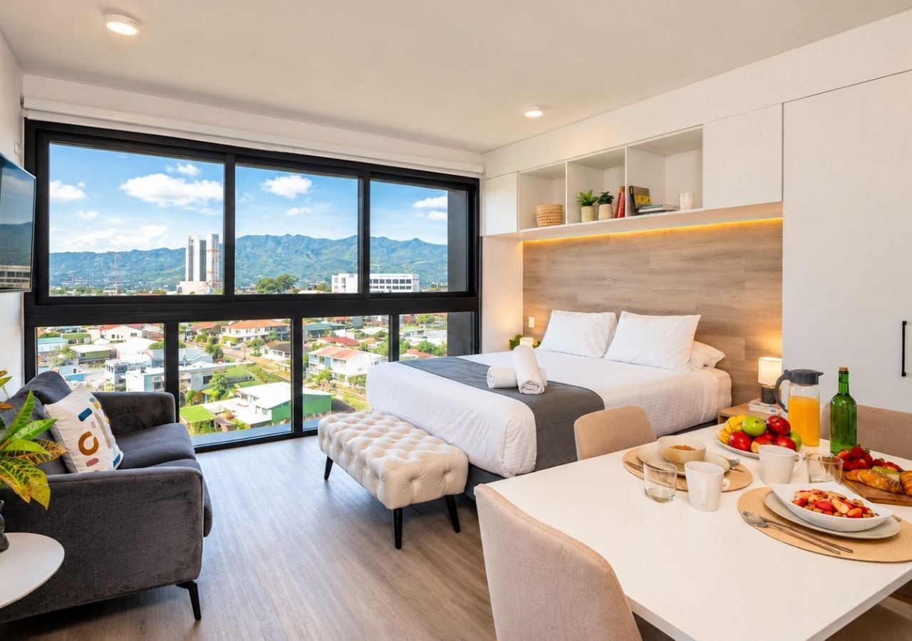 Une chambre d’hôtel moderne dotée d’une grande fenêtre, d’un lit, d’un canapé, d’une table à manger où le petit-déjeuner est servi, et offrant une vue sur la ville.