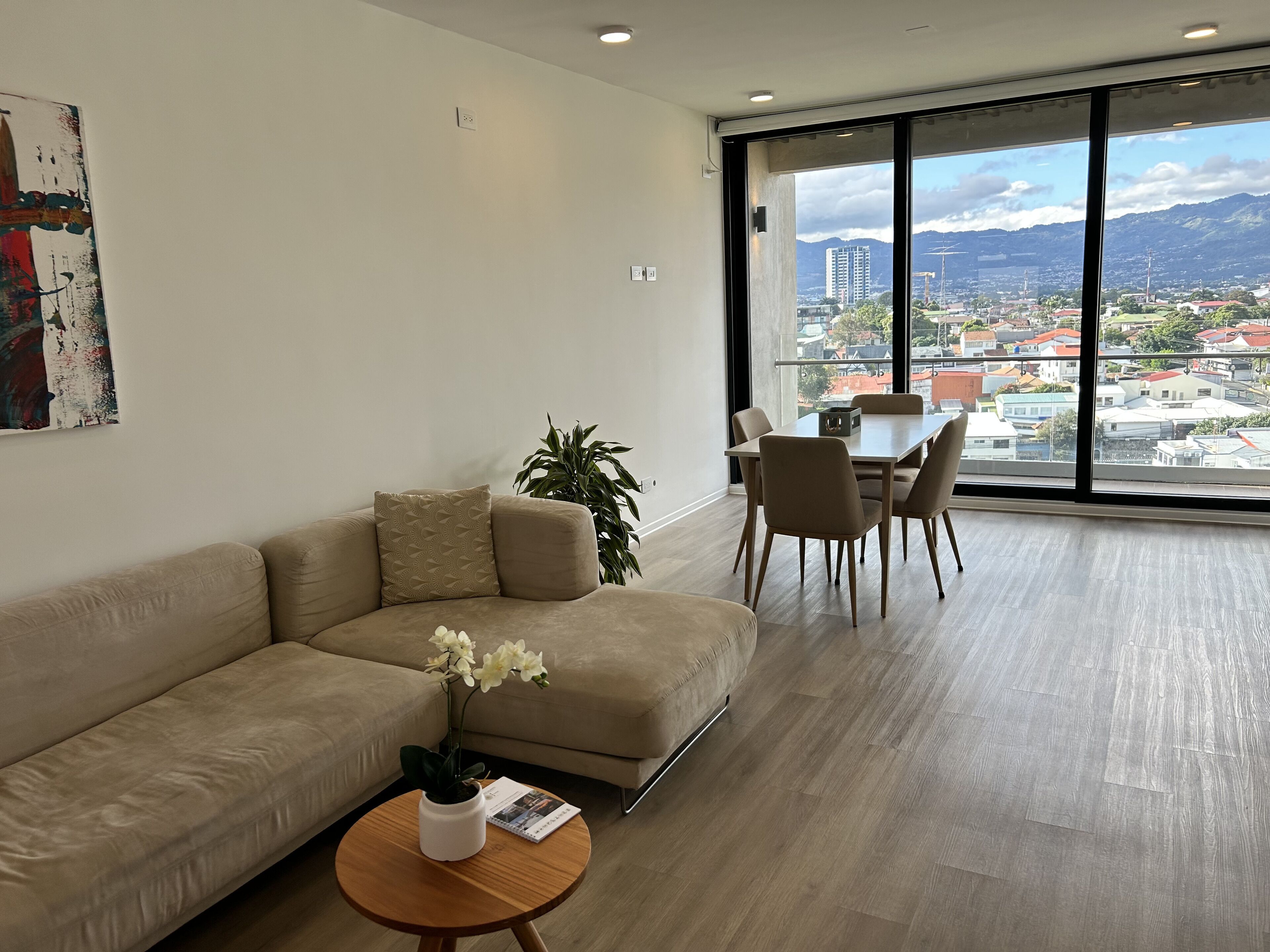 A modern living room with a beige sofa, a dining table with chairs, a potted plant, and a view of a cityscape through large windows.