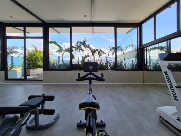 Une salle de sport moderne dotée de grandes fenêtres, d’équipements d’exercice et offrant une vue sur des palmiers et des montagnes.