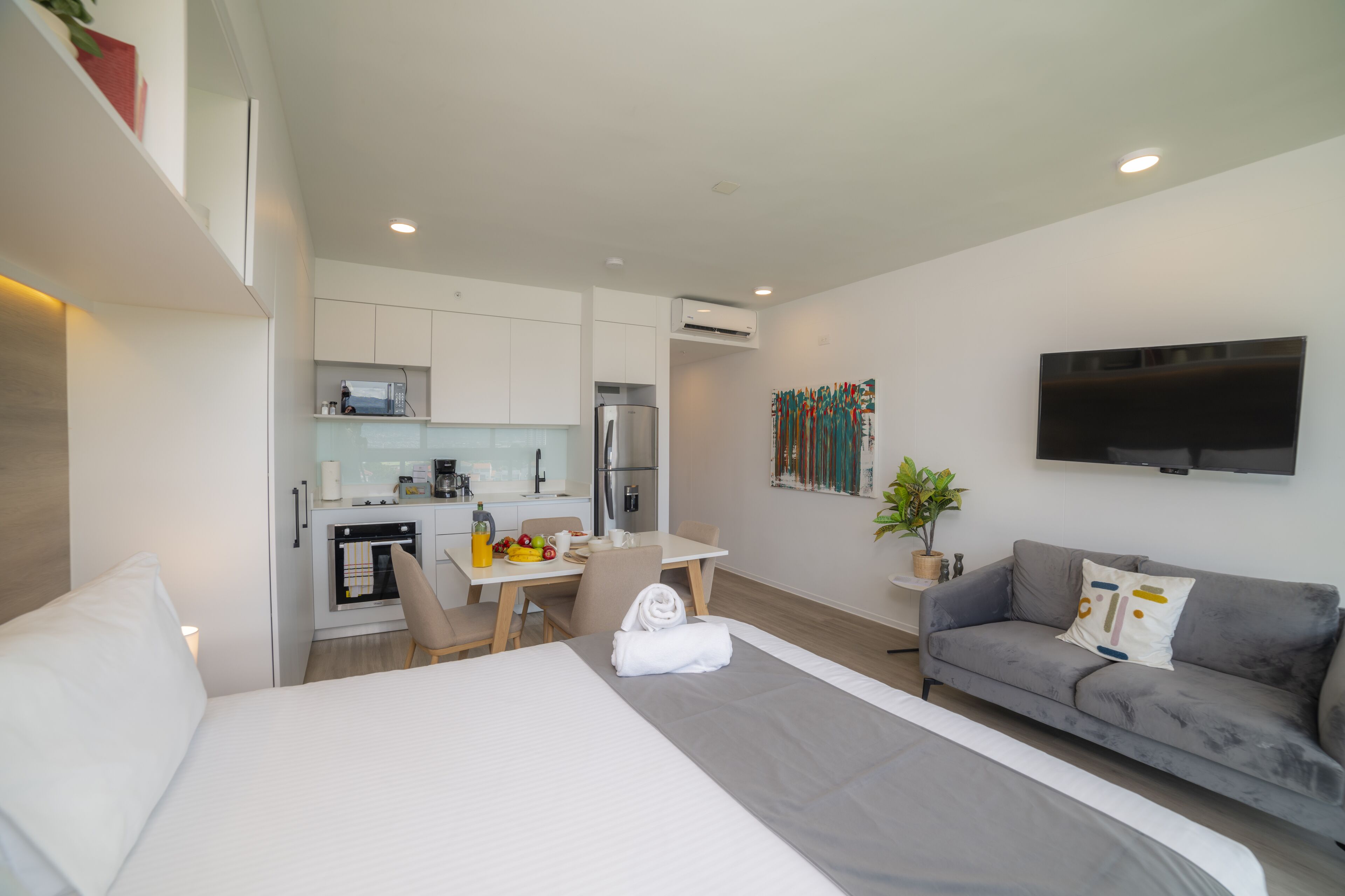 A modern hotel room with a bed, a grey sofa, a dining table with chairs, a kitchen area, and a flat-screen TV mounted on the wall.
