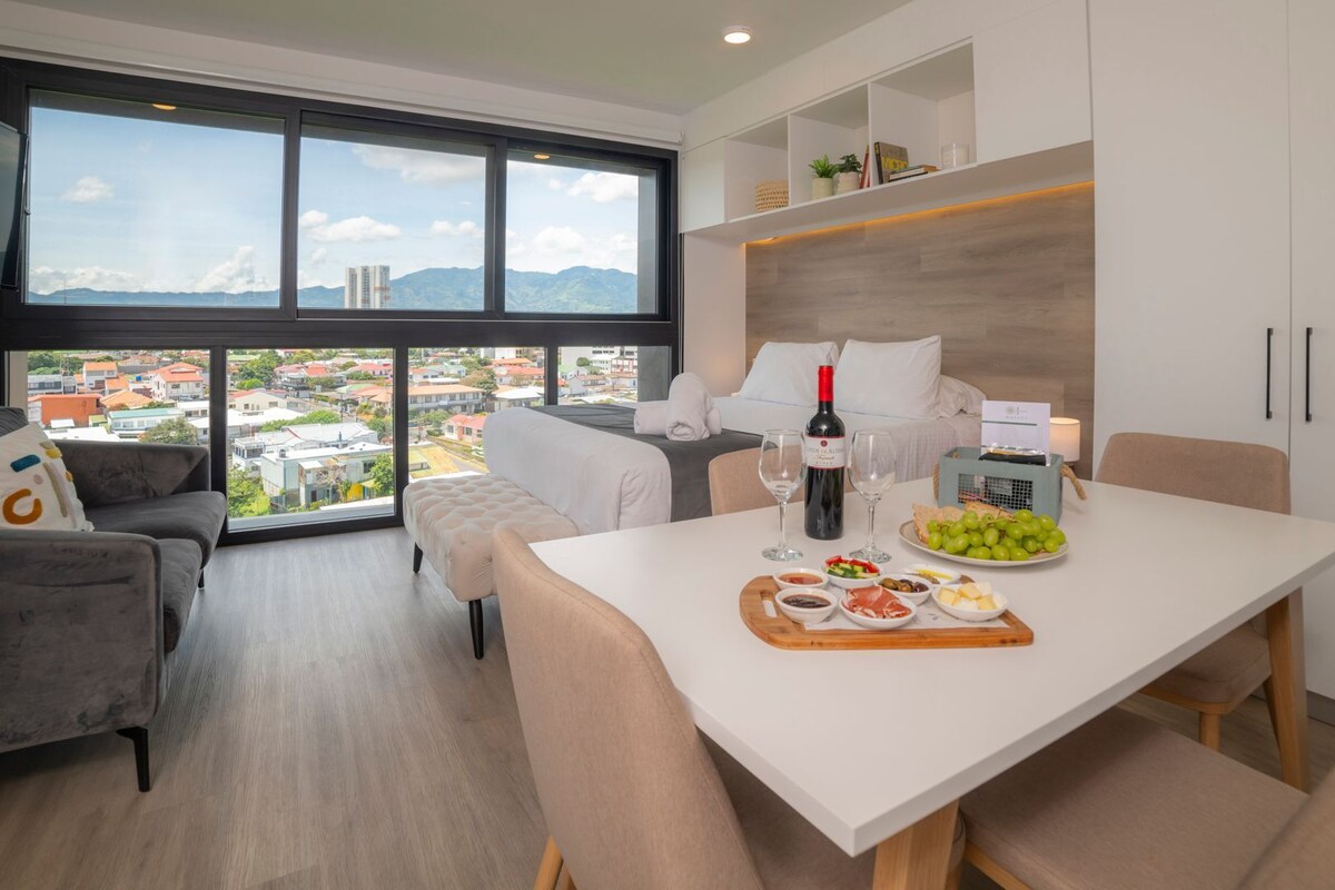 Une chambre d’hôtel moderne dotée d’une grande fenêtre donnant sur la ville, d’un coin repas avec une table dressée pour deux personnes, et d’un espace salon confortable avec un canapé et un fauteuil.