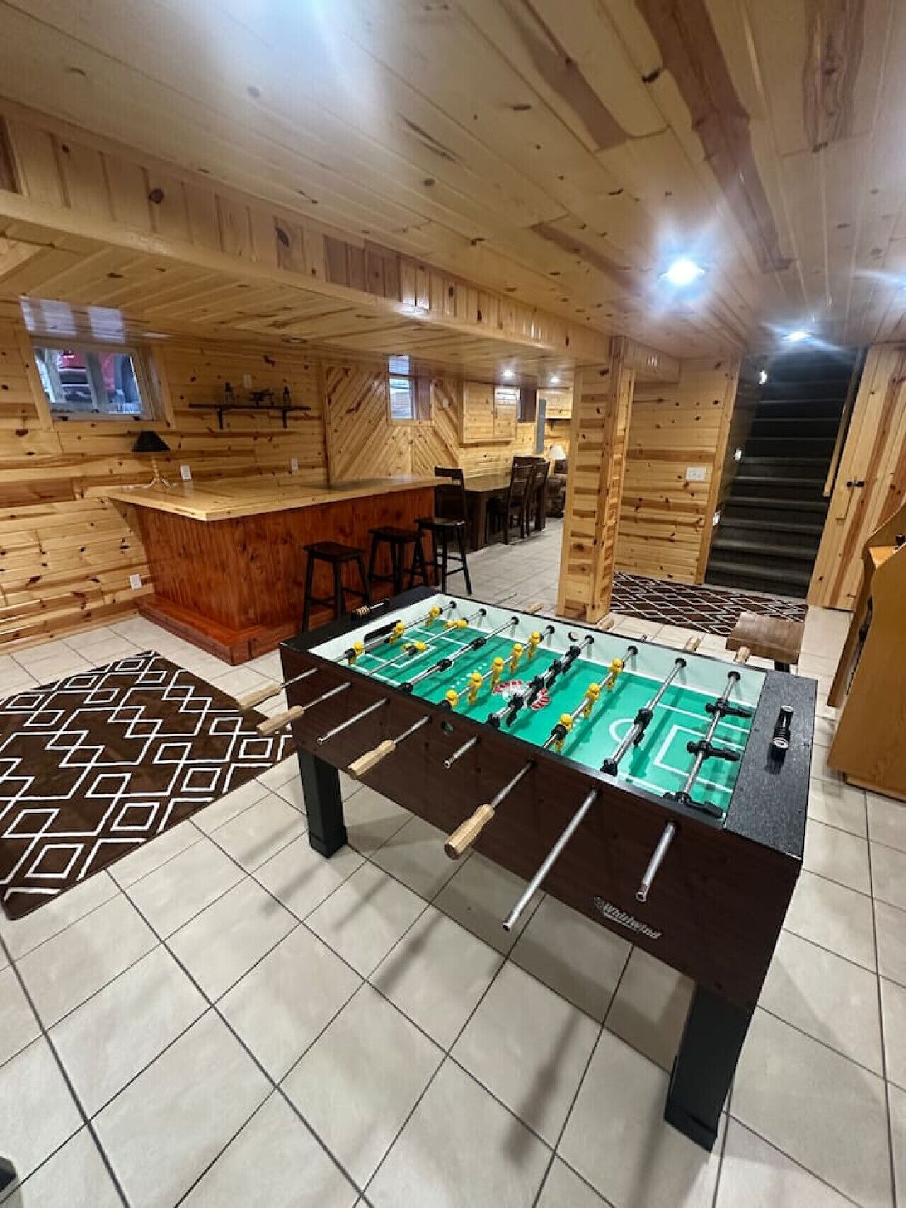 Una habitación con piso de madera, mesa de fútbol de mesa, zona de bar y una escalera.