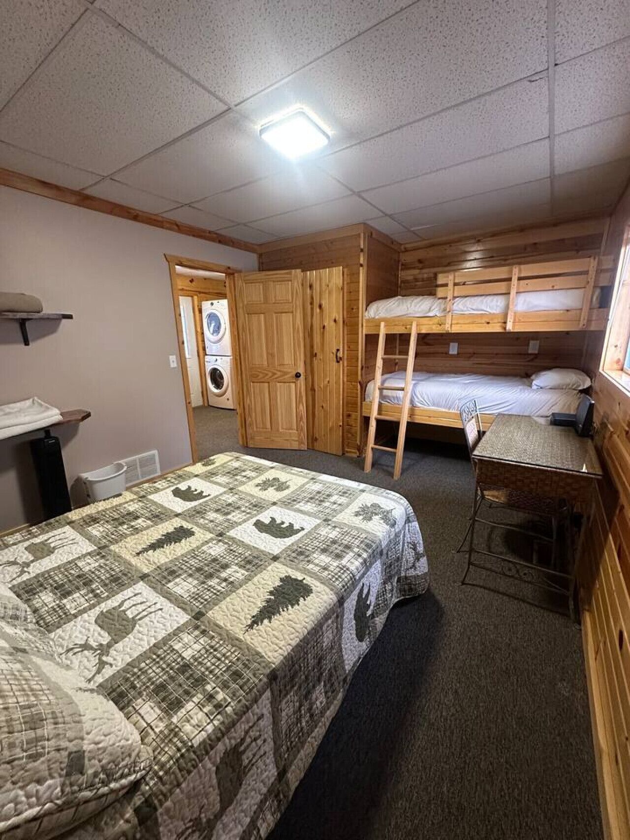 A small, compact room with bunk beds, a single bed with a quilt, a desk, and a washing machine in the corner.