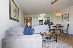 Un salon moderne avec un canapé gris, des coussins bleus, une table basse en verre et une table à manger en bois avec des chaises. On aperçoit une cuisine à l’arrière-plan, avec des meubles blancs et un luminaire suspendu.