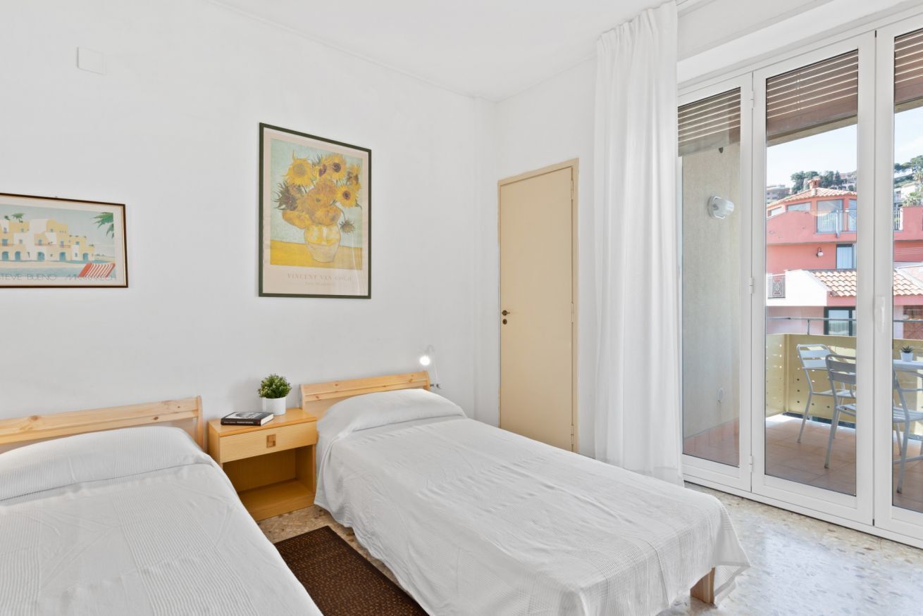 Una habitación con dos camas, una mesita de noche con lámpara, un cuadro en la pared y un ventanal corredero que da a un espacio exterior.