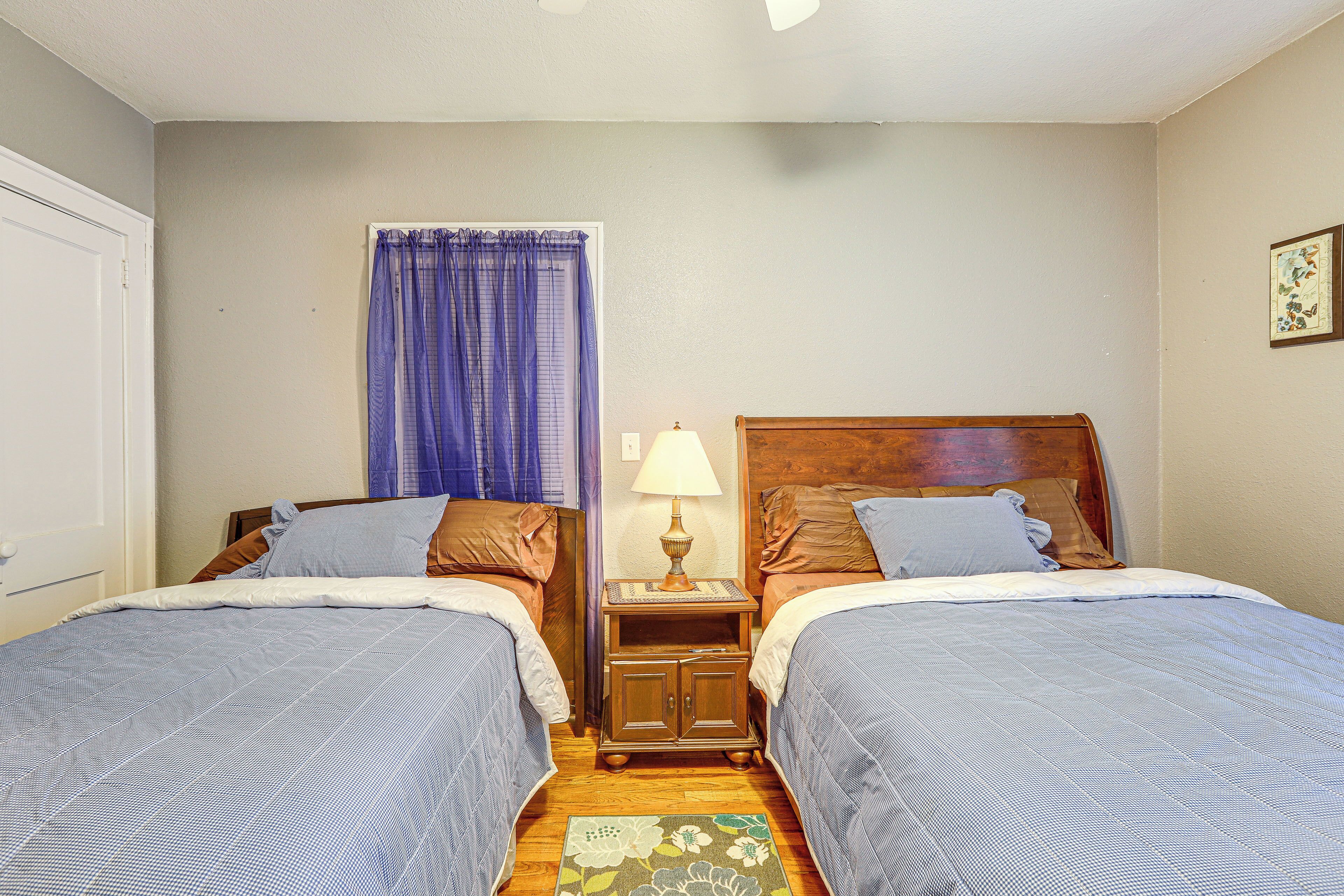Quarto duplo com duas camas, mesa de cabeceira com abajur, janela com cortinas e cabeceira de madeira.