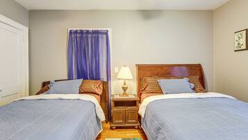 Quarto duplo com duas camas, mesa de cabeceira com abajur, janela com cortinas e cabeceira de madeira.
